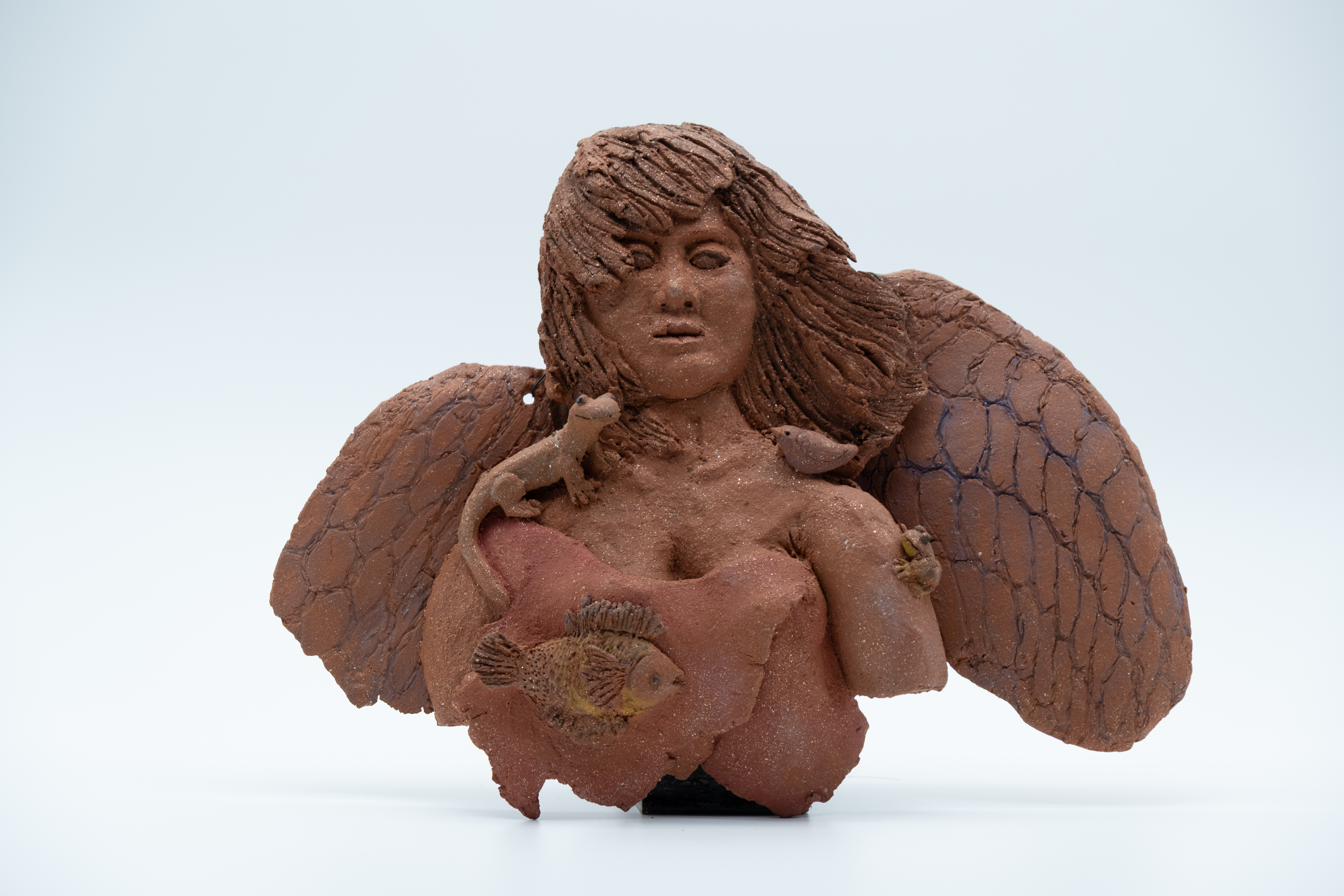 Clay bust of a winged angel figure with long flowing hair, textured wings, and small creatures including a lizard, a bird, and a fish resting against her chest