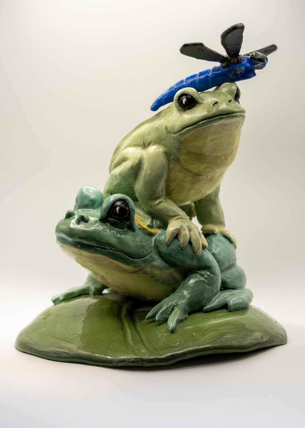 Clay sculpture of two glazed green frogs on a lily pad with a blue dragonfly perched on the upper frog's head