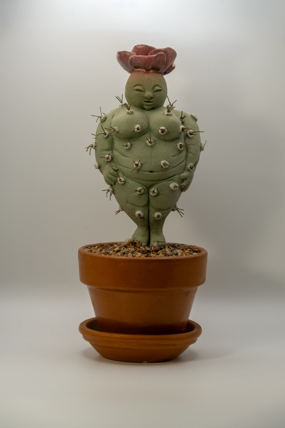 Whimsical mixed-media sculpture of a rounded green cactus goddess figure studded with spines and topped with a pink succulent bloom, planted in a terracotta pot