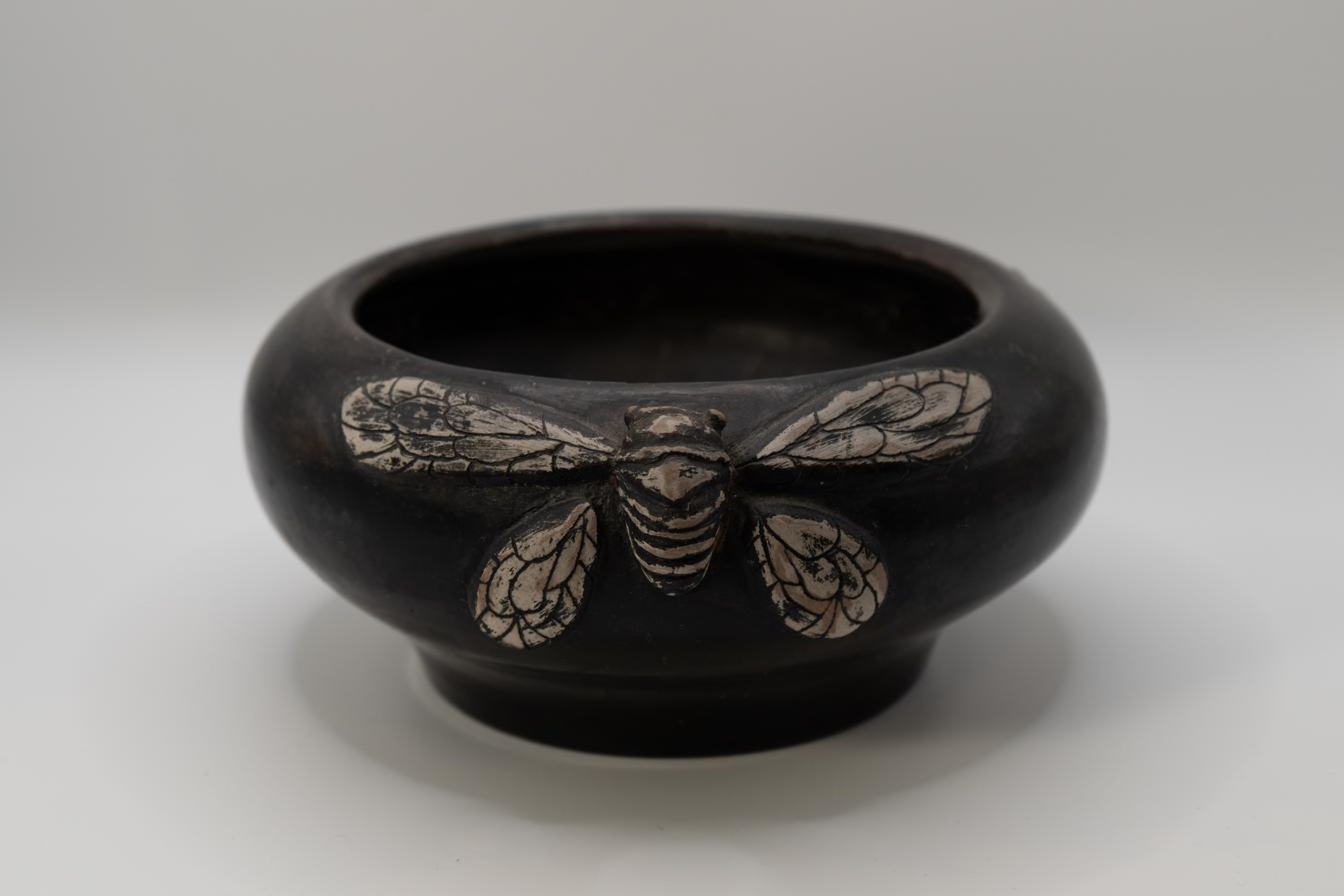 Dark pit-fired terracotta bowl with a cicada sculpted and painted in pale tones across its rim, wings outstretched on either side