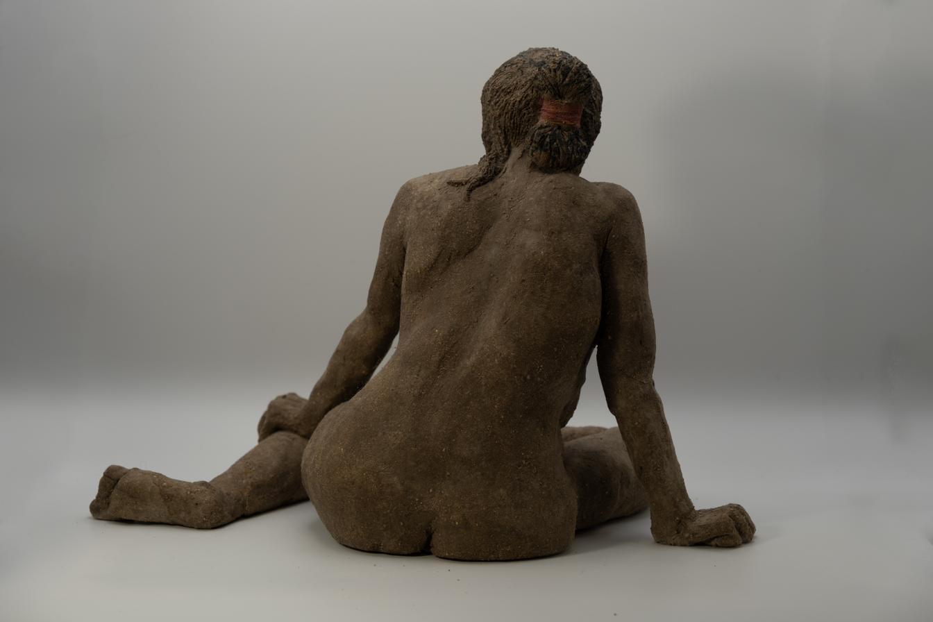 Air-dried clay sculpture of a seated nude figure viewed from behind, long fiber hair and beard bound with a cord, knees drawn to one side