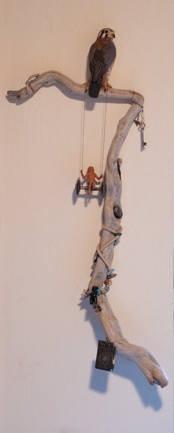 Mixed-media assemblage of a sculpted American kestrel perched on a tall piece of driftwood hung with a small clay figure on a swing, an antique key, a locket, and found beads and stones