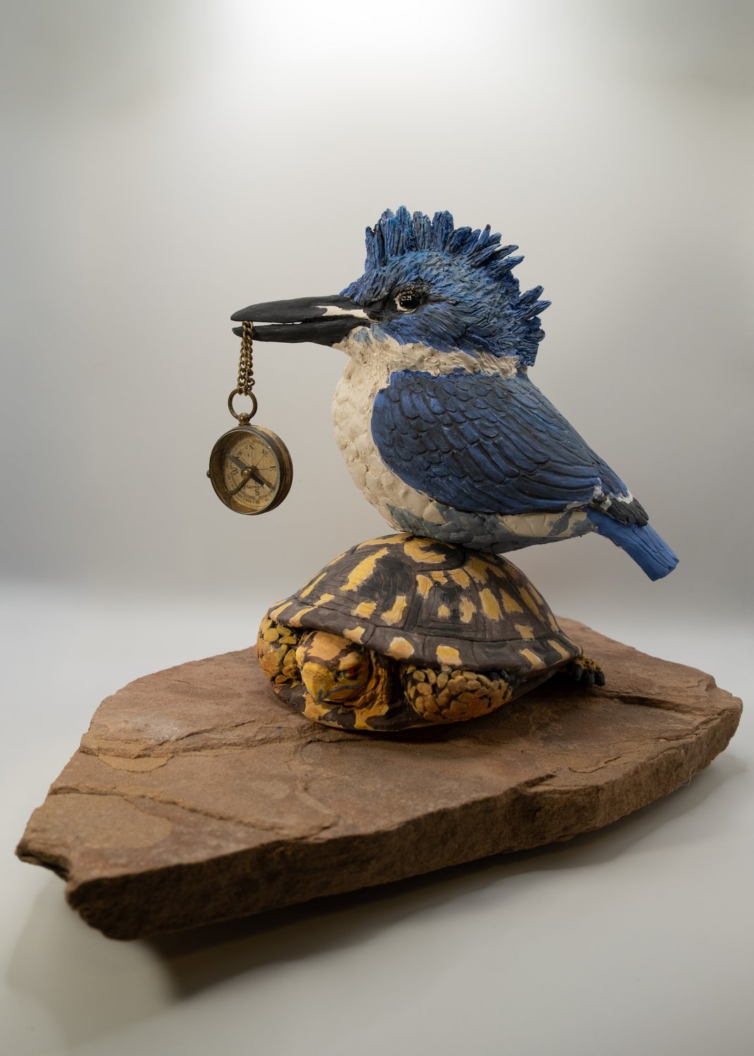 Clay sculpture of a belted kingfisher holding a vintage brass compass on a chain in its beak, perched on a box turtle atop a flagstone base