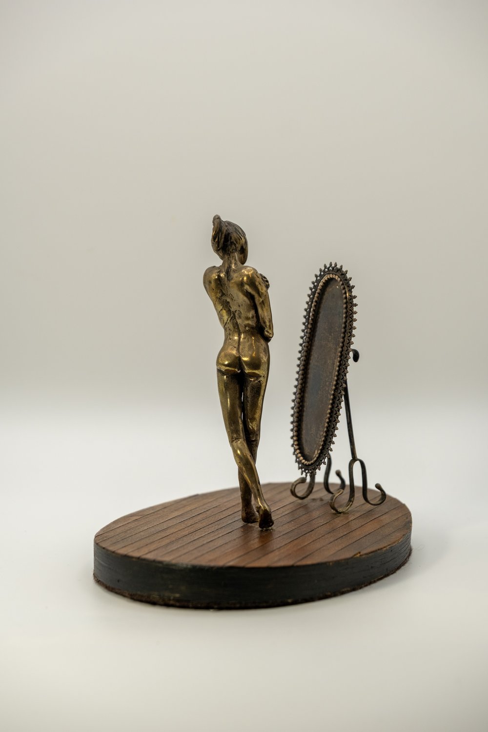 Mixed-media sculpture of a cast metal figure of a young woman standing before a small oval mirror on an oval wooden base, viewed from behind