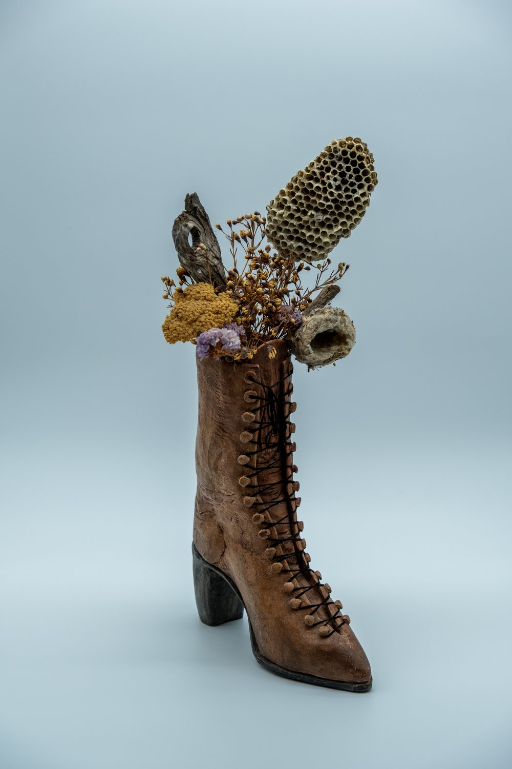 Mixed-media sculpture of a ceramic Victorian lace-up boot holding an arrangement of dried flowers, driftwood, and a papery wasp nest
